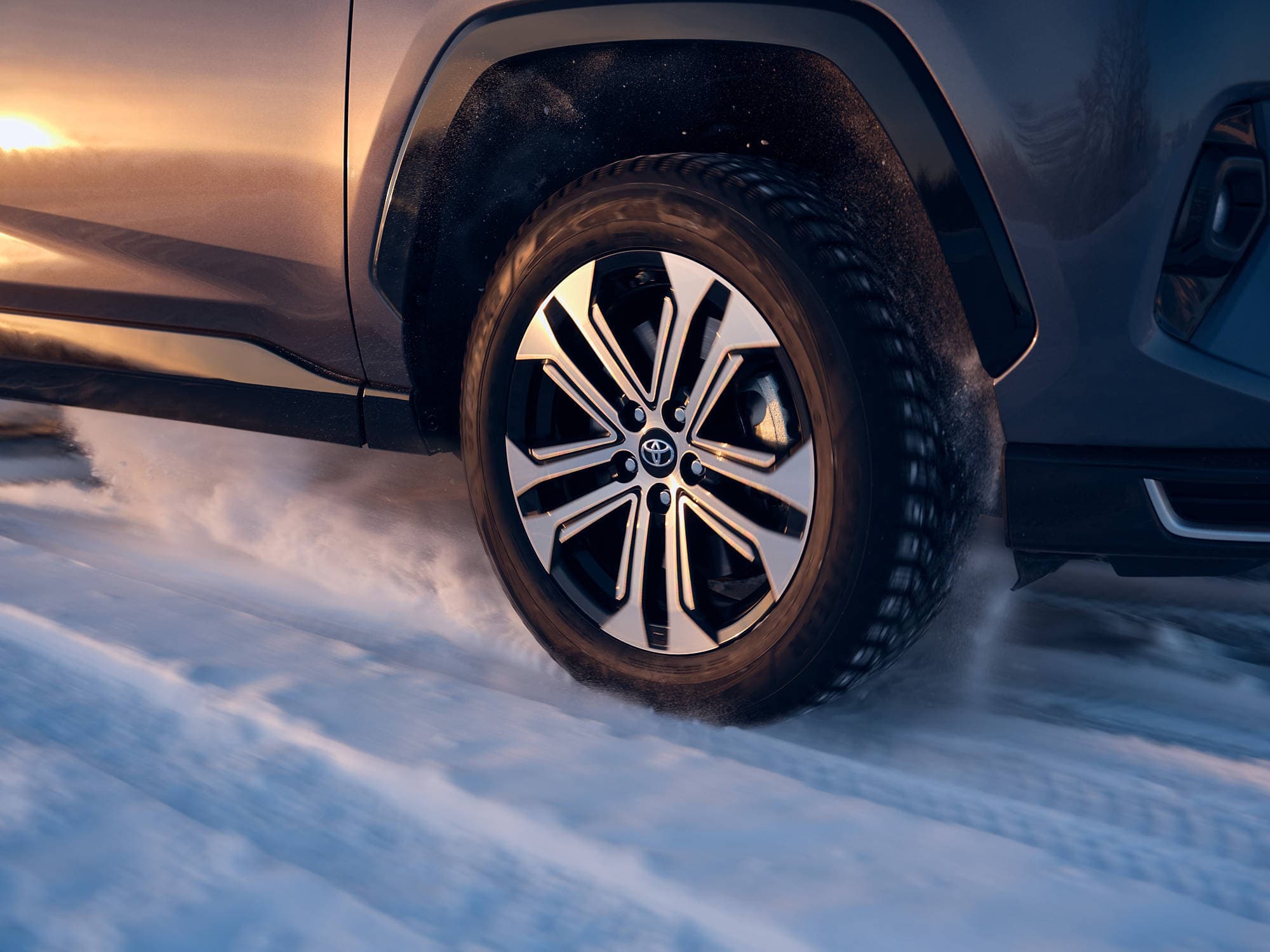 Close-up of a car tire driving through snow, with snow powder being kicked up, and a sunset reflecting on the vehicle's side.