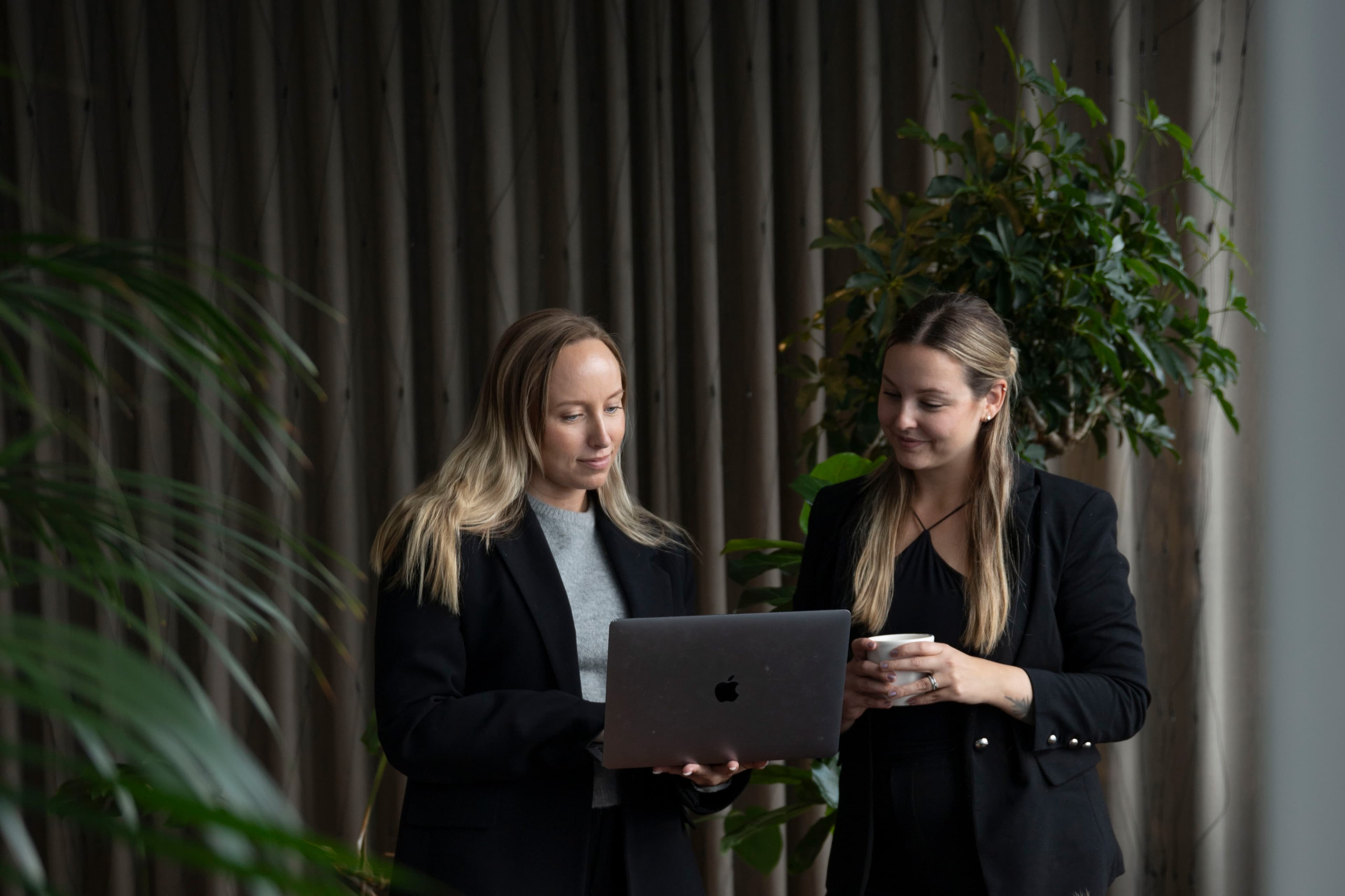 Server-side tracking Two women in black attire are looking at a laptop screen. One holds a mug. They stand in a softly lit room with plants in the background.